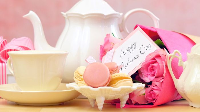 A white teapot and matching cup next to a plate of macarons and a bouquet of roses, with a card reading "Happy Mother's Day" in fancy cursive script.
